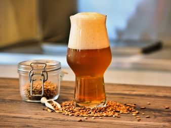 Belgisches Bier in tulpenförmigem Glas mit dichter Schaumkrone auf Holztisch, umgeben von verstreutem Getreide und Glasgefäß mit Getreide.