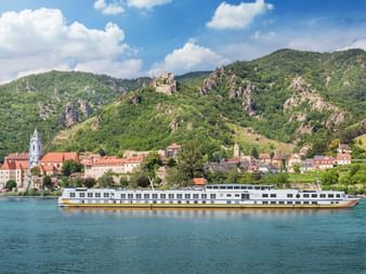 White river cruise ship MS Vista Classica sailing on the Danube with a historic town, church towers, and green mountains with castle ruins in background.