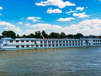 White and blue MS Switzerland river cruise ship sailing on calm water under a bright blue sky with white clouds. Trees and buildings visible on shore.
