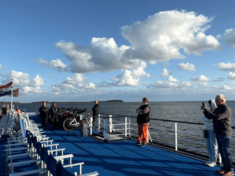Passagiere auf blauem Fährdeck bei Überfahrt nach Rügen. Menschen stehen an Reling und fotografieren Meerblick unter teilweise bewölktem Himmel.