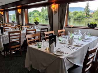Elegant dining room aboard the Classica ship with white-clothed tables, wooden chairs, and large windows overlooking a river and green landscape.