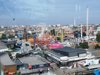 Luftaufnahme des Wiener Praters mit dem ikonischen Riesenrad, bunten Fahrgeschäften und umliegendem Stadtbild unter blauem Himmel.