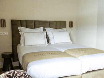 Modernes Hotelzimmer mit Doppelbett, weißer Bettwäsche, beigem Läufer, gepolstertem Kopfteil und bronzefarbenen Wandleuchten in der Magnifique X Junior Suite.