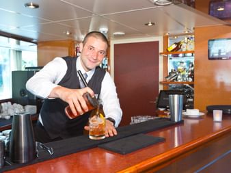Smiling bartender in vest and white shirt pouring whiskey into glass at wooden bar counter on MS SE-MANON cruise ship.