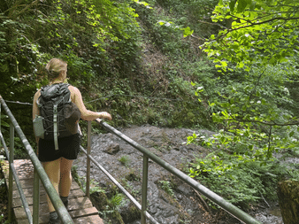 Wanderin mit Rucksack überquert Holzsteg mit Metallgeländer in der Ruppertstklamm, umgeben von üppiger grüner Vegetation und Felswänden.