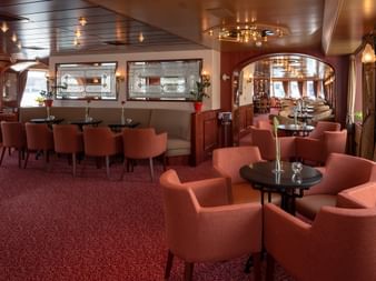 Elegant salon aboard the Switzerland ship with coral-colored armchairs, round tables, red carpet, and wood-paneled ceiling with chandeliers.