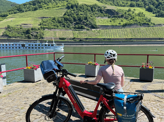 Radfahrerin mit rotem E-Bike blickt auf Rhein und Schiff Olympia, mit terrassierten Weinbergen an Hängen im Hintergrund.