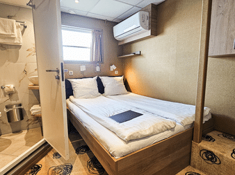 Kompakte Oberdeckkabine auf MS Nassau mit Doppelbett, weißer Bettwäsche, eigenem Bad, Fenster, Klimaanlage und gemustertem Teppich.
