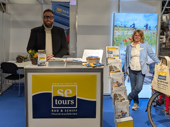 Two people at se-tours booth: man in black suit behind counter, woman in blue jacket beside brochure display. Bicycle wheel and scenic backdrop visible.