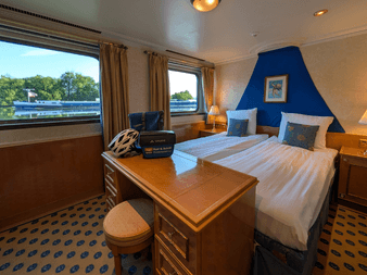 Geräumige Kabine mit Doppelbett, blauer Akzentwand, Holzschreibtisch, gemustertem Teppich und großen Fenstern mit Flussblick auf der Casanova.