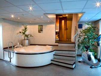Moderner Wellnessbereich auf MS SE-MANON mit rundem Whirlpool, Holzsauna, Heimtrainer und grünen Pflanzen unter Einbauleuchten.