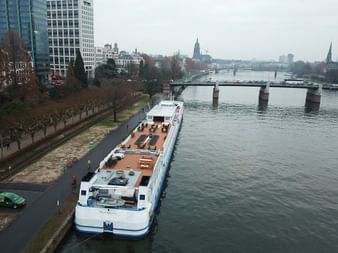 White river cruise ship MS Classica docked at urban riverbank with bridges and city skyline in background under overcast sky.