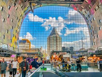 Innenansicht der Rotterdamer Markthalle mit bunter Deckenmalerei, Essensständen und Besuchern. Die Bogendecke zeigt blauen Himmel mit Wolken.
