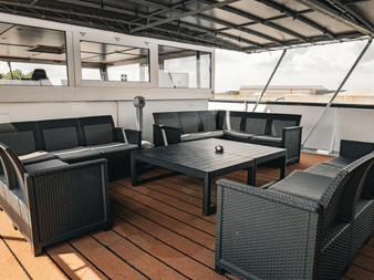 Sonnendeck der MS De Nassau mit schwarzen Rattan-Loungemöbeln um einen Tisch auf Holzdeck unter einem überdachten Bereich.