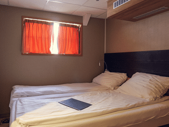 Kompakte Unterdeck-Kabine auf Nassau mit Doppelbett, dunklem Kopfteil, beigen Wänden, orangefarbenem Vorhang und Holzdecke.