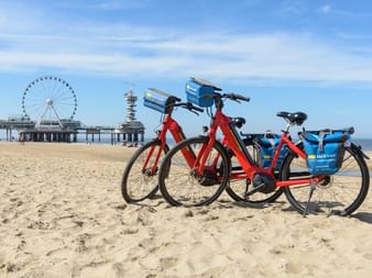 Zwei Leihfahrräder am Sandstrand von Scheveningen mit Pier und Riesenrad im Hintergrund unter blauem Himmel mit Wolken.
