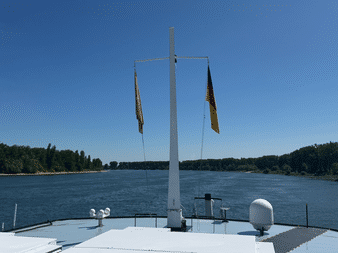 Blick vom Schiffsbug mit deutscher Flagge am Mast, fahrend auf breitem Fluss mit grünen bewaldeten Ufern unter klarem blauen Himmel.