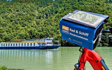Blau-weißes Flusskreuzfahrtschiff auf grünem Wasser vor bewaldeten Hügeln. Rotes Fahrrad mit blauer SE-Tours Tasche im Vordergrund.