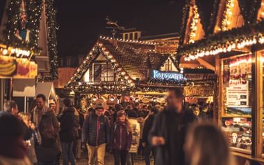 Evening scene at a Christmas market with wooden stalls decorated with warm lights. Visitors walk between illuminated booths selling food and gifts.