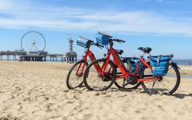 Zwei Leihfahrräder am Sandstrand von Scheveningen mit Pier und Riesenrad im Hintergrund unter blauem Himmel mit Wolken.