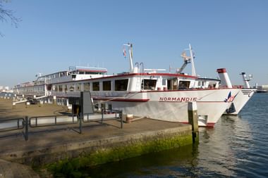 Weißes Passagierschiff MS Normandie mit rotem Zierstreifen am Betonpier vertäut. Das Schiff hat mehrere Decks mit großen Fenstern.