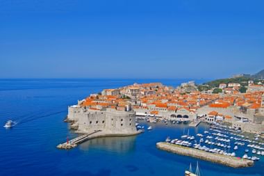 Dubrovniks Altstadt und Hafen von oben Luftaufnahme von Dubrovniks ummauerte Altstadt mit roten Ziegeldächern, Steinbefestigungen und Marina voller weißer Boote im blauen Wasser.