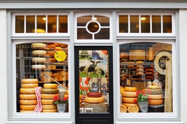 Storefront of a cheese shop in Gouda with large windows displaying stacks of yellow cheese wheels and Dutch-themed decorations.