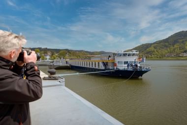 Fotograf mit grauen Haaren fotografiert Flusskreuzfahrtschiff Prinzessin Katharina am Kai der Donau bei Wien mit Hügeln im Hintergrund.