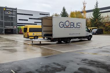 Weißer Globus Lieferwagen vor moderner Parkhaus-Einfahrt. Gelber Bus im Hintergrund. Nasser Asphalt und Herbstbäume sichtbar.