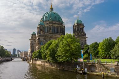 Berlin Cathedral (Berliner Dom) with distinctive green copper domes beside the Spree River, surrounded by lush green trees under a cloudy sky.