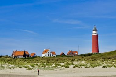 Leuchtturm Texel mit Dorfhäusern Rot-weißer Leuchtturm Texel steht hoch hinter einer Reihe traditioneller niederländischer Häuser mit orangen Ziegeldächern, umgeben von Dünen.