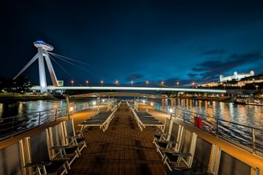 Nachtansicht vom Sonnendeck der MS Serenade 1 mit beleuchteter Brücke, Stadtlichtern im Wasser und Liegestühlen auf Holzboden.