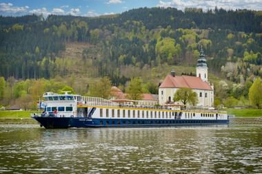 MS Prinzessin Katharina Flusskreuzfahrtschiff fährt auf ruhigem Wasser mit weißer Kirche und bewaldeten Hügeln im Hintergrund unter blauem Himmel.