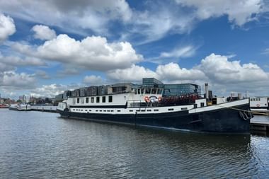 Weißes Passagierschiff Anna van Aemstel mit dunklem Rumpf am Kai vertäut. Moderne Gebäude im Hintergrund unter bewölktem blauen Himmel.