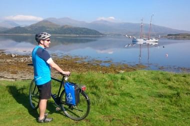 Radfahrer in blauem Shirt mit Helm steht neben Fahrrad am grasbewachsenen Ufer und blickt auf ruhigen See mit Flying Dutchman Segelschiff.