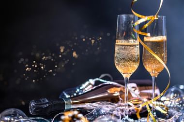 Zwei Sektgläser mit Sekt vor dunklem Hintergrund mit goldenem Band, Sektflasche und festlicher Dekoration.