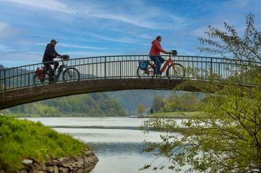 Zwei Radfahrer mit Fahrrädern überqueren eine geschwungene Fußgängerbrücke über die Donau, umgeben von grüner Vegetation und Hügeln.