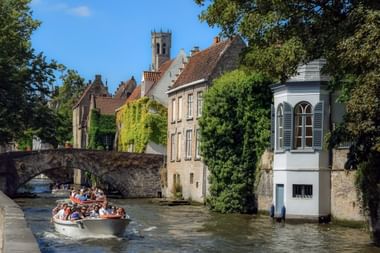 Touristenboot mit Passagieren auf einem Kanal in Brügge, unter einer Steinbrücke mit mittelalterlichen Gebäuden und Kirchturm im Hintergrund.
