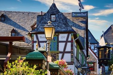Traditionelle Fachwerkhäuser in Rüdesheims Drosselgasse mit bunten Fassaden, Schieferdächern und Blumenschmuck unter blauem Himmel.