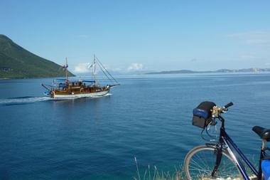Traditionelles Holzfischerboot Panagiota segelt auf ruhigem blauen Wasser mit Bergen im Hintergrund. Blaues Fahrrad mit Taschen am Ufer geparkt.