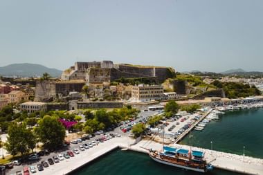 Luftaufnahme des Korfu Hafens mit türkisfarbenem Wasser, Booten und der Alten Festung auf einem Hügel über der Mittelmeerküste.