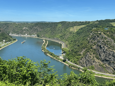 Luftaufnahme der Loreley-Felsformation und Rheinschleife mit Frachtschiff, Bahnlinie und bewaldeten Hängen unter blauem Himmel.