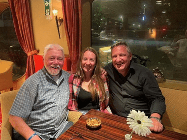 Three smiling people seated at a restaurant table with red curtains and windows. A white flower and small bowl are on the wooden table.