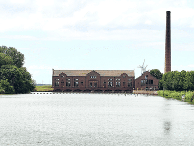 Historisches rotes Backsteingebäude des Woudagemaal-Pumpwerks mit hohem Schornstein neben einem Kanal in Lemmer. Windturbine im Hintergrund.