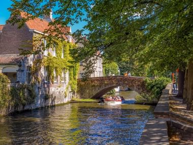 Steinbrücke über Kanal in Brügge mit efeubedeckten mittelalterlichen Gebäuden, Touristenboot darunter und baumgesäumtem Gehweg an sonnigem Tag.