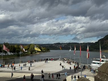 Wide plaza at Deutsches Eck in Koblenz with visitors, flags, and river confluence. Cloudy sky above, hills and buildings in background.