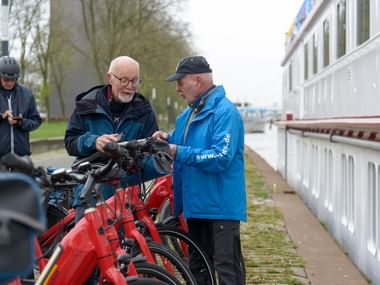 Zwei Männer betrachten rote Leihräder auf einem Weg neben einem weißen Flusskreuzfahrtschiff. Weitere Radfahrer sind im Hintergrund bei Bäumen sichtbar.