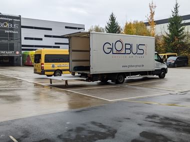 White Globus service van parked at entrance of modern parking garage. Yellow bus visible in background. Wet pavement and autumn trees visible.