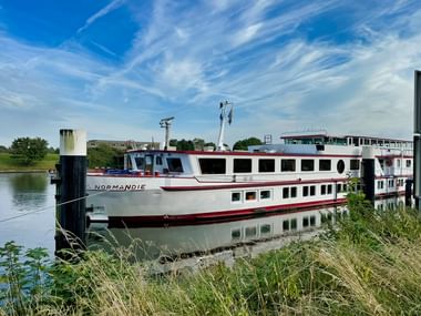 Weißes Flusskreuzfahrtschiff MS Normandie mit rotem Zierstreifen am Anleger eines ruhigen Flusses, umgeben von grüner Vegetation unter blauem Himmel.