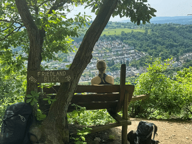Person sitzt auf Holzbank am Aussichtspunkt Friedland Blick mit Blick auf ein Tal mit Stadt und Fluss, umgeben von bewaldeten Hügeln.